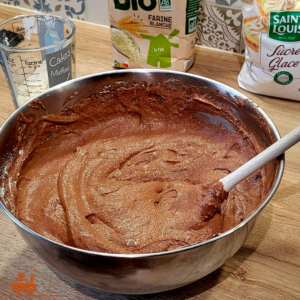 Un appareil à génoise au chocolat dans un saladier avec une cuillère en bois. En arrière plan un verre doseur contenant de la farine, un paquet de farine et un paquet de sucre glace.