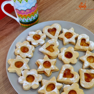 Assiette grise contenant des biscuits Linzer Augen de différentes formes (étoiles, coeurs, fleurs) à la confiture d'abricots et tasse à thé rose avec des motifs