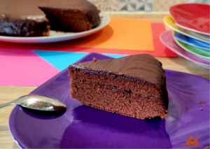 Une part de gâteau Sacher (sachertorte) posée sur une assiette violette avec une cuillère à café retournée. En arrière plan une sachertorte et une pile d'assiettes colorées.