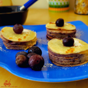 une assiette bleue contenant 5 myrtilles et 3 mini-gâteaux de crêpes à la confiture de myrtilles décorés avec des myrtilles et du sucre glace