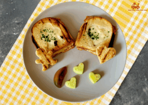 Nappe de bistrot jaune sous une assiette grise contenant 2 croque-monsieur dont le dessus est taillé à l'emporte-pièce pour faire des motifs : un bonhomme et un cœur accompagnés d'une tranche de tomate et de 3 morceaux de concombre taillés en forme de cœur.
