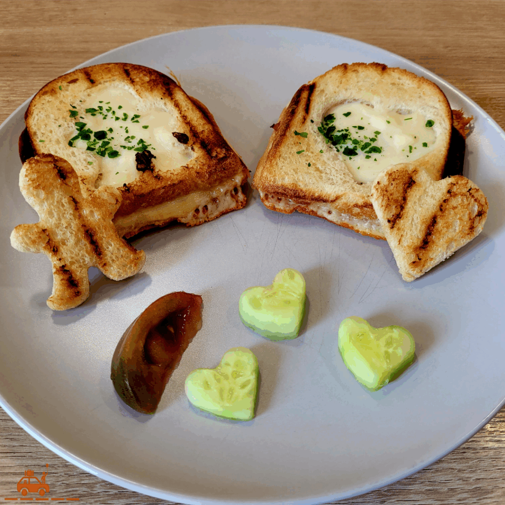 Une assiette grise contenant 2 croque-monsieur dont le dessus a été taillé à l'emporte-pièce et 2 morceaux de pain de mie en forme de cœur et de bonhomme, une tranche de tomate et 3 morceaux de concombre en forme de cœur.