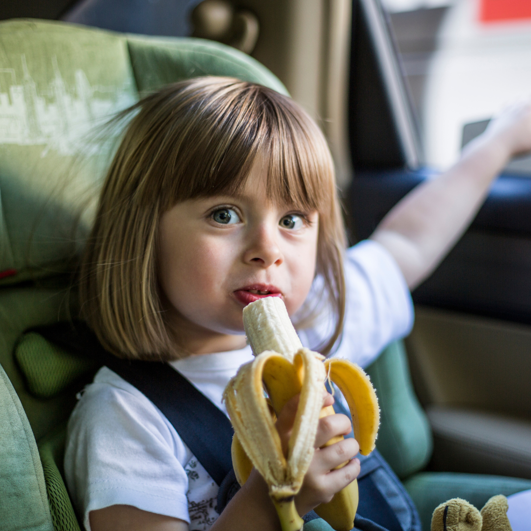 Petite fille occupée à manger une banane dans la voiture : leur faire manger des snacks est une bonne idée pour occuper les enfants dans la voiture quand ils s'ennuient