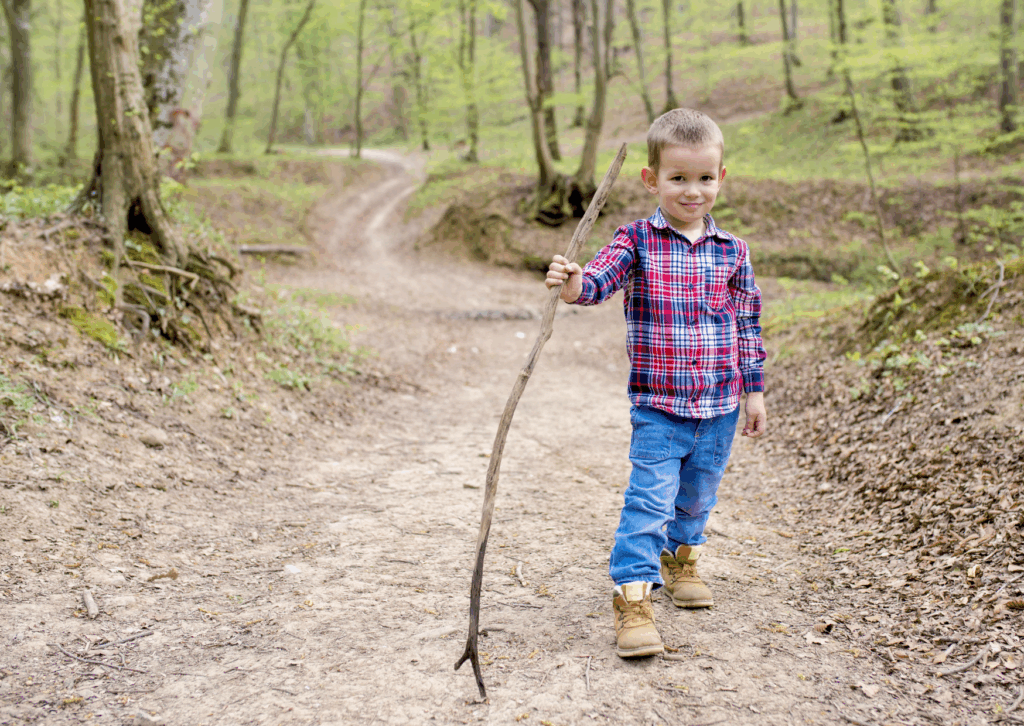 Enfant de 5 ans avec des chaussures de randonnées sur un sentier de forêt qui tient un grand bâton dans la main