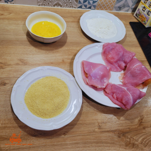 4 cordons bleus maison garnis à l'italienne avec de la mozzarella et du jambon de parme et posés sur une assiette et 3 assiettes contenant de l'oeuf cru battu, de la farine blanche et de la chapelure pour préparer la panure du cordon bleu