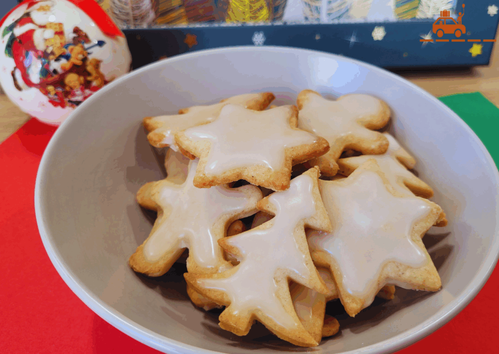 Bol de biscuits de Noël à la cannelle avec un glaçage blanc au sucre glace. Les biscuits sont en forme de bonshommes, d'étoiles et de sapins. En arrière plan, une boule de Noël et une boîte décorée avec des étoiles.