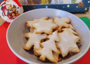 Bol de biscuits de Noël à la cannelle avec un glaçage blanc au sucre glace. Les biscuits sont en forme de bonshommes, d'étoiles et de sapins. En arrière plan, une boule de Noël et une boîte décorée avec des étoiles.