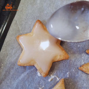 Un biscuit en forme d'étoile posé sur une plaque recouverte de papier sulfurisé et recouvert d'un glaçage blanc, une petite cuillère entrain d'étaler le glaçage sur le biscuit. C'est la dernière étape de la recette alsacienne de bredele à la cannelle. 