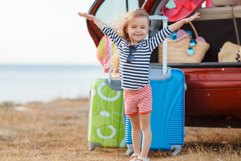 Petite fille souriante devant le coffre d’une voiture rempli de valises avant un voyage en famille