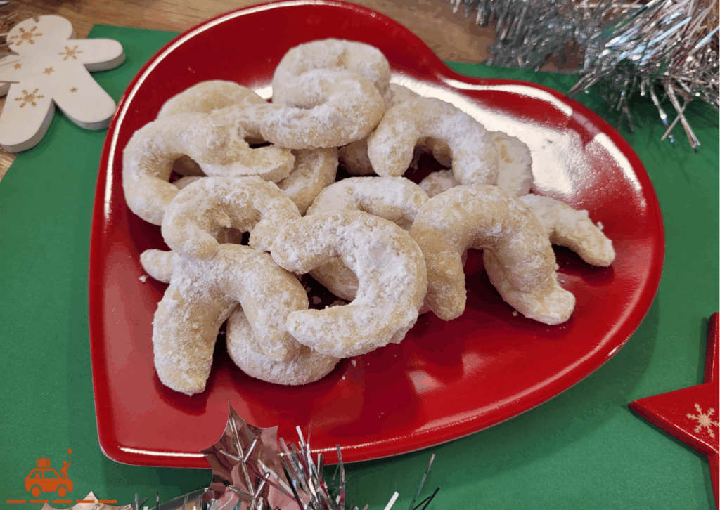 Une assiette rouge contenant des Vanille Kipferl : des biscuits autrichiens en forme de croissant recouverts de sucre glace et parfumés à la vanille. Le tout entouré de décorations de Noël
