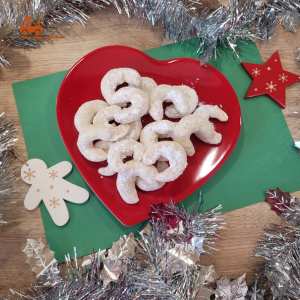Des décorations de Noël entourant une assiette rouge en forme de coeur contenant des Vanille Kipferl : des biscuits de Noël autrichiens recouverts de sucre glace et en forme de croissants